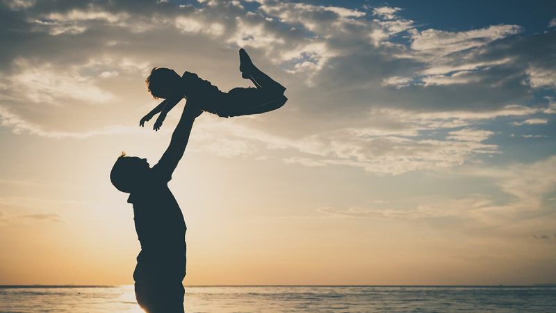 man holding son above head on beach in sunset